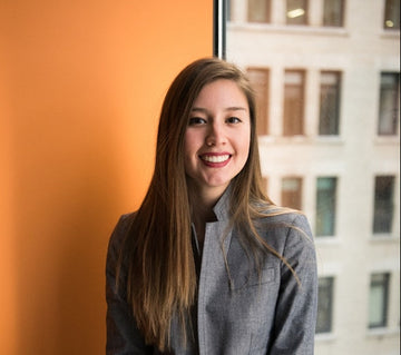 Woman in a gray blazer sitting by a window with an orange wall.